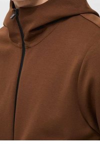 Felpa con cappuccio marrone caratterizzata da un colletto alto e una cerniera nera. Realizzata in un tessuto morbido con una superficie liscia. Design pulito con accenti minimi.