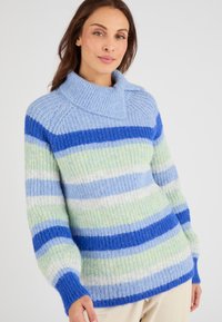 Maglione a righe in tonalità di blu, verde e bianco con una texture spessa e lavorata a maglia, e un alto collo a coste. Maniche lunghe con polsini blu.