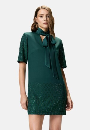 Donna che indossa un vestito a maniche corte verde scuro con dettagli di perline a motivo di diamante e una sciarpa annodata al collo, in piedi davanti a uno sfondo neutro.