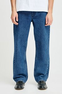 Jeans de mezclilla azul con un corte holgado, piernas rectas, textura ligeramente deslavada y detalles de costura, combinados con zapatos negros brillantes.