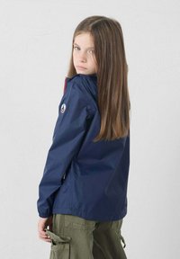 Jongen meisje met lang steil haar, gekleed in een marineblauwe jas en olijfgroene cargobroek, die over haar schouder kijkt tegen een effen achtergrond.