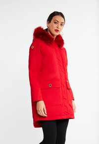 Cappotto invernale rosso con cappuccio in pelliccia sintetica, chiusura con zip, due tasche frontali e orlo posteriore più lungo. Tessuto liscio e dettagli di cucitura tono su tono.