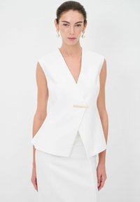 Blazer bianco senza maniche con scollatura a V profonda, design strutturato e chiusura con dettagli in oro. Tessuto liscio, vestibilità sartoriale, estetica minimalista.
