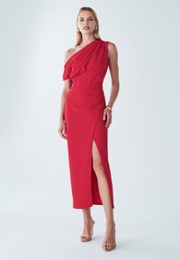 Robe rouge à une épaule avec un corsage ajusté, détails drapés et une fente haute sur la cuisse. Tissu lisse et silhouette élégante. Talons hauts transparents visibles.