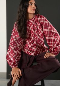 Kvinde siddende iført en rød satinbluse med hvid skotskternet og lange, puffede ærmer, kombineret med mørke breddebenede bukser og sorte hæle.