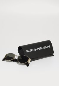 Ochelari de soare cu formă rotundă de pisică, cu lentile întunecate, accente metalice aurii, așezati lângă un carcasă neagră cu text alb "RETROSUPERFUTURE".