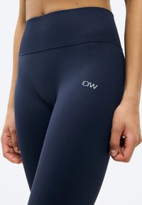 Κοντινή λήψη ενός ατόμου που φοράει σκούρες μπλε κολάν με ψηλή μέση, ραβδωτή μέση και μικρό ανακλαστικό λογότυπο "ICIW" στο μηρό.