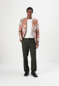 Blazer mit floralem Muster in Weiß mit rosa und grünen Akzenten, kombiniert mit olivgrünen Hosen und schwarzen Schuhen. Das Model schaut direkt in die Kamera.
