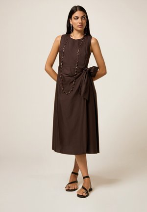 Braunes ärmelloses Kleid mit Taillenzug, das eine fließende Silhouette und glattes Material aufweist. Getragen mit schwarzen Sandalen und einer Perlenkette.