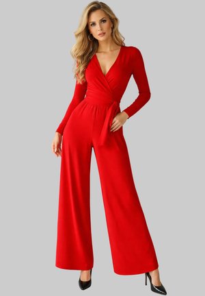 Donna con lunghi capelli biondi che indossa una tuta rossa a maniche lunghe con scollo a incrocio e tacchi alti neri, in piedi davanti a uno sfondo grigio uniforme.