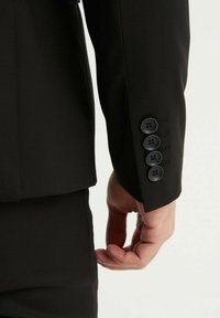 Manche de veste noire à boutons noirs, tissu texturé et coupe ajustée avec un léger rétrécissement à l'extrémité.