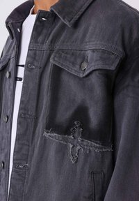 Giacca in denim nero con finitura testurizzata, presenta una piccola toppa strappata, due tasche frontali e chiusure a pulsante a scatto.