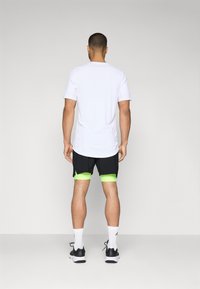 T-shirt de sport blanc, shorts noirs avec bordure vert néon, chaussures de sport noires, chaussettes blanches mi-mollet ; vue de dos.