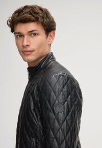 Giovane uomo con capelli ricci che indossa una giacca in pelle trapuntata nera con cerniera e una toppa rotonda sul maniche, rivolto leggermente verso sinistra.