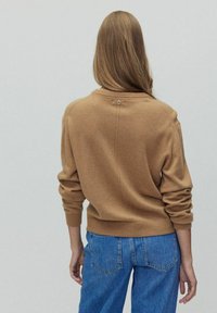 Pull en tricot marron avec col rond, poignets côtelés et détail de boutons au dos, associé à un jean en denim bleu.