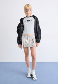 Grijze crop top met zwarte accenten, zwarte oversized jas, gestreepte grijze geplooide minirok en witte sneakers met grijze details.