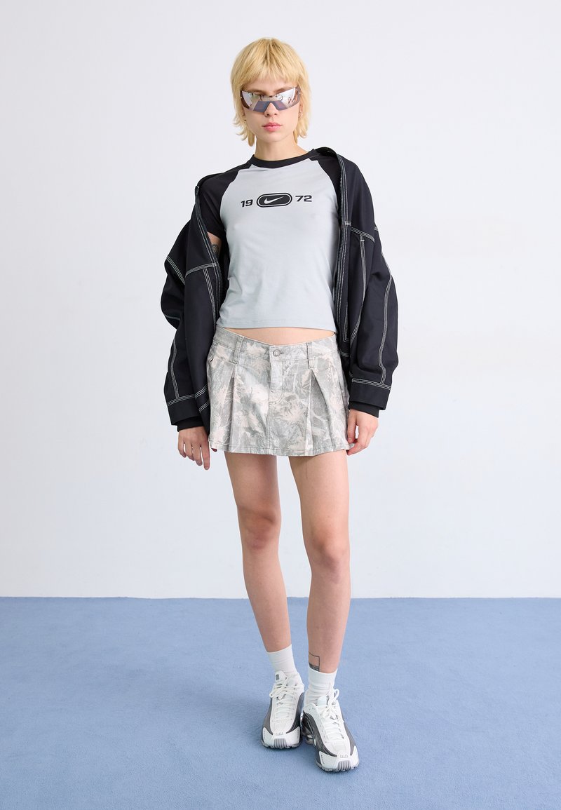 Top corto gris con acentos negros, chaqueta negra oversized, falda mini plisada gris con estampado, y zapatillas blancas con detalles grises.
