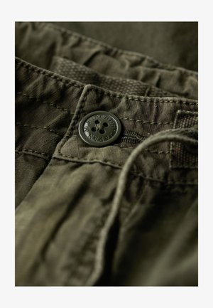 Olivengrøn bomuldsstof med en subtil tekstur, prydet med en rund sort knap præget med "SUPERDRY" nær linningen.