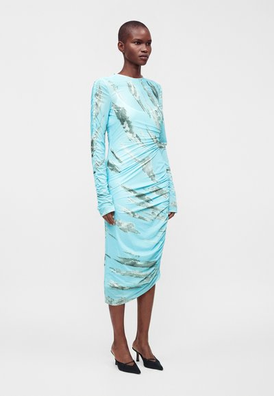 Femme aux cheveux courts portant une robe midi bleu clair à manches longues avec des motifs abstraits gris et des talons hauts pointus noirs.