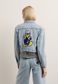 Lyseblå denimjakke med en tegneseriefigur brodert på baksiden. Figuren har solbriller og sort antrekk, med gule detaljer.