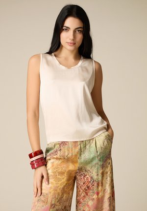 Młoda kobieta o długich ciemnych włosach, ubrana w kremowy, bezrękawnik, wielokolorowe, wzorzyste spodnie z szerokimi nogawkami oraz czerwone, warstwowe bransoletki.