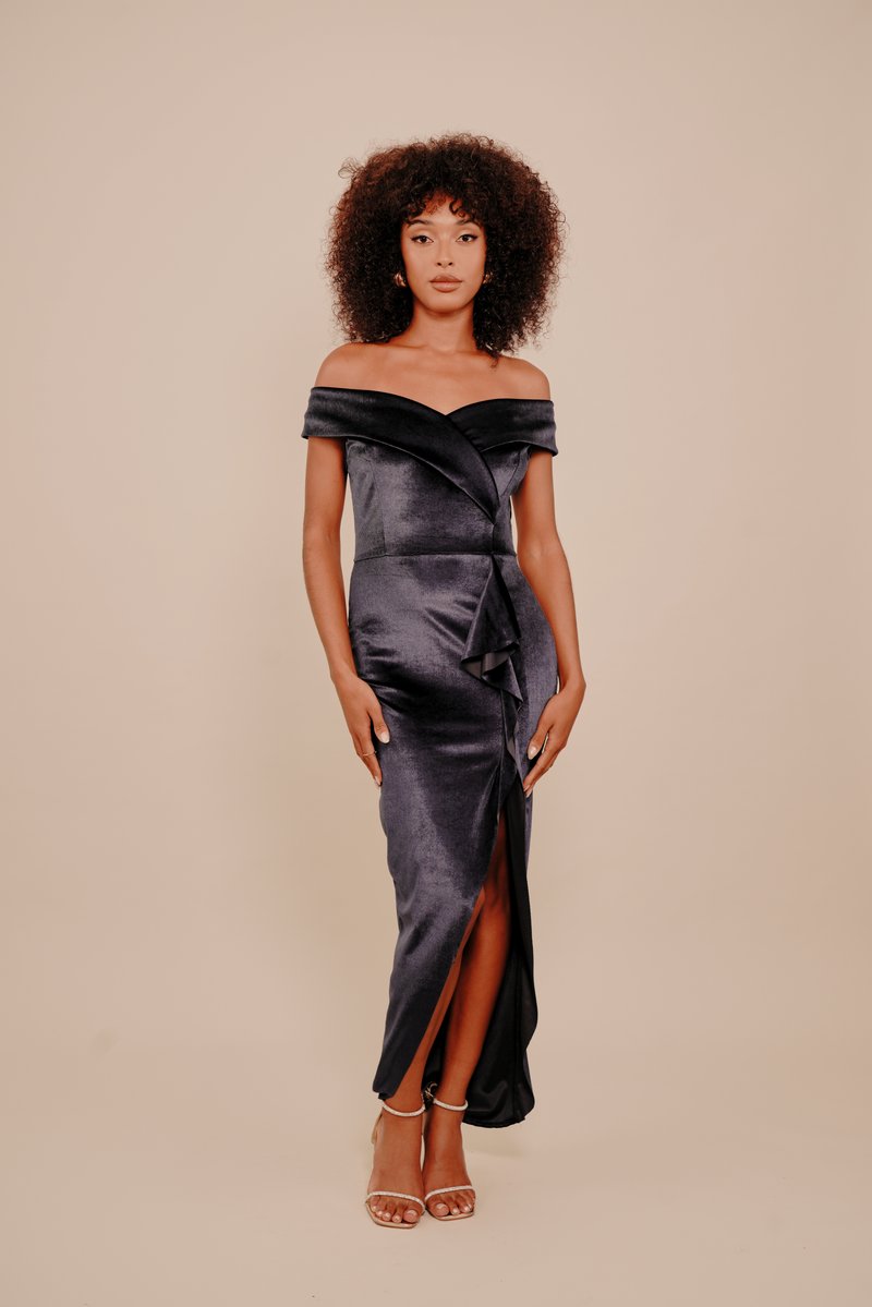 Göndör hajú nő fekete, váll nélküli, testhez simuló szatén estélyi ruhában, magas hasítékkal a combnál és bézs pántos magassarkúban, bézs háttér előtt áll.