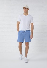 Shorts in cotone blu con cintura elastica e cordino, abbinati a una maglietta bianca semplice e sneaker bianche, davanti a un muro di mattoni bianchi.