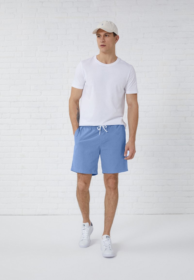 Shorts in cotone blu con cintura elastica e cordino, abbinati a una maglietta bianca semplice e sneaker bianche, davanti a un muro di mattoni bianchi.
