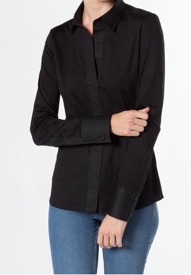 Femme portant une chemise noire ajustée à manches longues avec des poignets boutonnés et un jean bleu, debout avec un bras croisé sur l'autre.