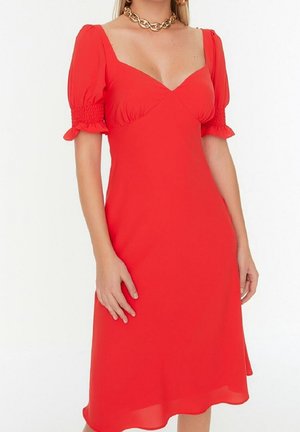 Vrouw draagt een rood jurkje tot op de knie met pofmouwen en een vierkante halslijn, geaccentueerd met een stevige gouden ketting.