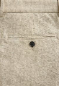 Tissu texturé beige avec une poche passepoilée unique munie d'une fermeture par bouton noir près du centre.