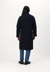 Cappotto blu navy in misto lana con uno spacco sul retro, abbinato a pantaloni in denim e scarpe nere. Design semplice e lineare.