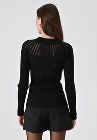 Maglione nero a costine con maniche lunghe, caratterizzato da un design con pannello trasparente sulla parte superiore della schiena. Indossato con pantaloni corti neri.