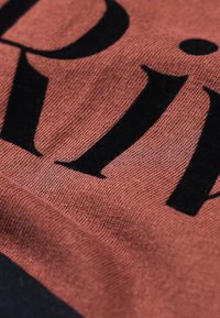 Pamuk tkanina u boji hrđe s velikim crnim tekstom. Tekstura je glatka s laganim sjajem, ističući detalje ispisa.