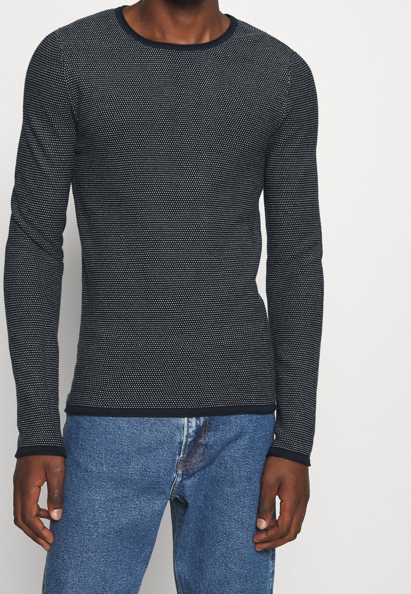 T-shirt à manches longues bleu marine avec un motif à pois texturé, col rond et bord noir contrastant, associé à un jean en denim bleu.