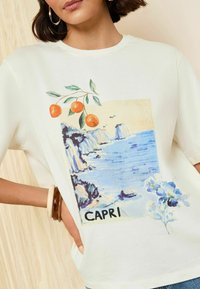 Balts T-krekls ar krāsainu grafiku, kas attēlo piekrastes ainavu ar klintīm, zilu ūdeni, oranžiem augļiem un vārdu "CAPRI."