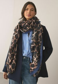Rutev šal s leopardjim vzorcem v toplih tonih, z raztrganimi robovi. Mehka tekstura, velika velikost, nošen čez modro srajco in črno blazer jakno. Priložnostni slog.