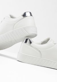Sneakers bianchi con tomaia in pelle liscia, suola rigata di un bianco grigiastro e dettagli argentati sul tallone. Presentano un design con lacci.
