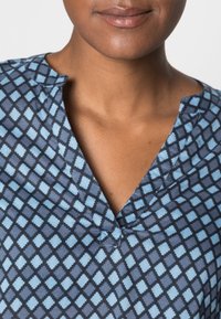 Blue and black patterned blouse with a V-neck, featuring a geometric design of diamonds in varying shades of blue and subtle collar details.