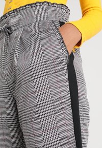 Pantalons à carreaux noir et blanc avec une taille froncée, des poches latérales et une bande latérale contrastante noire. Fabriqués en tissu texturé.