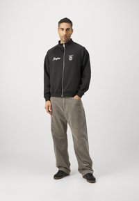 Sort zip-up jakke med hvidt logo og tekst, parret med lysegrå kordfløjl bukser og sorte sneakers. Stående på en ensfarvet baggrund.