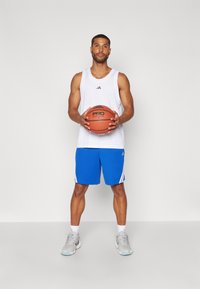 Hvit tanktopp med svart logo, blå shorts med hvite striper, holder en brun basketball, står på en lys grå bakgrunn.