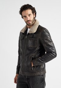 Zwarte leren jas met een beige nepbont kraag, ritssluiting aan de voorkant en twee borstzakken, met een gladde textuur en een getailleerd ontwerp.