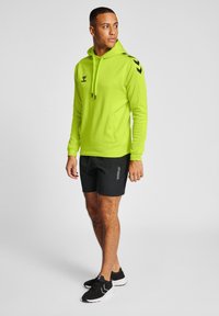 Limegrüner Pullover mit Kapuze, schwarzen Akzenten, Fronttasche und Kordelzug. Kombiniert mit schwarzen Shorts und schwarzen Sportschuhen.