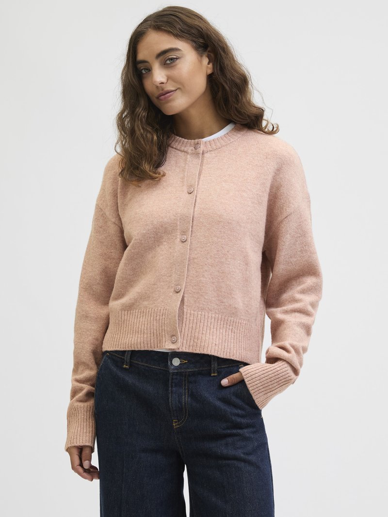 Ung kvinne med bølgete brunt hår som har på seg en lys rosa knappet cardigan og mørkeblå jeans, stående mot en ensfarget lys bakgrunn.