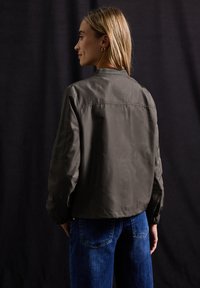 Grön bomberjacka med texturerad yta, elastiska ärmar och avslappnad passform, kombinerad med blå denimjeans. Bakgrunden är mörk.