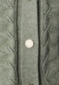 Neulejakku hillityn vihreänä, jossa on teksturoitu kuosi, sisältäen rivin metallinappeja ja koristeellista resoria reunoilla.