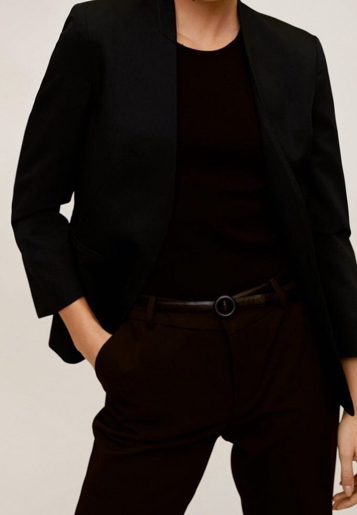 Personne portant un blazer noir ajusté, un haut noir et un pantalon noir avec une ceinture noire, debout avec une main dans la poche.