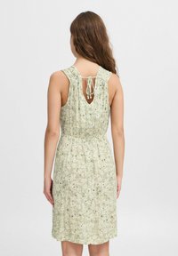 Robe florale vert clair avec un dos en scoop et un lien ajustable, dotée d'une taille froncée et d'un tissu texturé. Petits accents noirs sur l'ensemble.