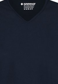 Donker marineblauwe V-hals shirt met het label "Garage Bio-Katoen Body Fit" en wasymbolen gedrukt aan de binnenkant van de kraag.